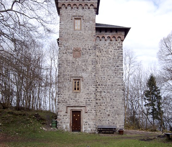 Een stenen toren met kantelen en een wapenschild, omringd door kale bomen. Op de voorgrond staan banken en een tafel op een weiland., © Alois Schneider