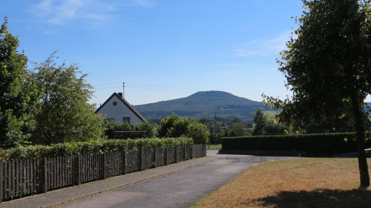 Eine ruhige Landschaft mit einem Weg, der zu einem Hügel führt. Im Vordergrund sind Bäume und ein Zaun zu sehen, unter einem klaren blauen Himmel.