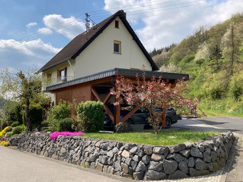 A charming house with a covered entrance and blooming plants. In the background, gentle hills are visible under a blue sky.