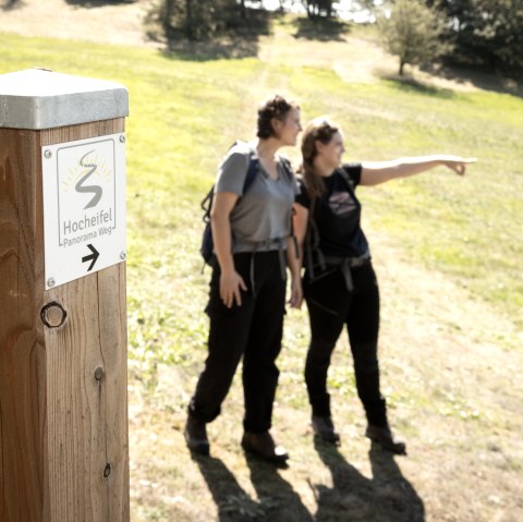 Zwei Wanderer stehen auf einem sonnigen Feldweg. Ein Holzpfosten zeigt den 'Hocheifel Panorama Weg' an. Eine Person zeigt in die Ferne., © TI Hocheifel-Nürburgirng©BaumannFotografie