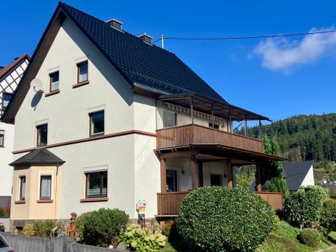 Une maison moderne avec un toit noir et un balcon. Entourée d'un jardin entretenu et d'un ciel bleu clair.