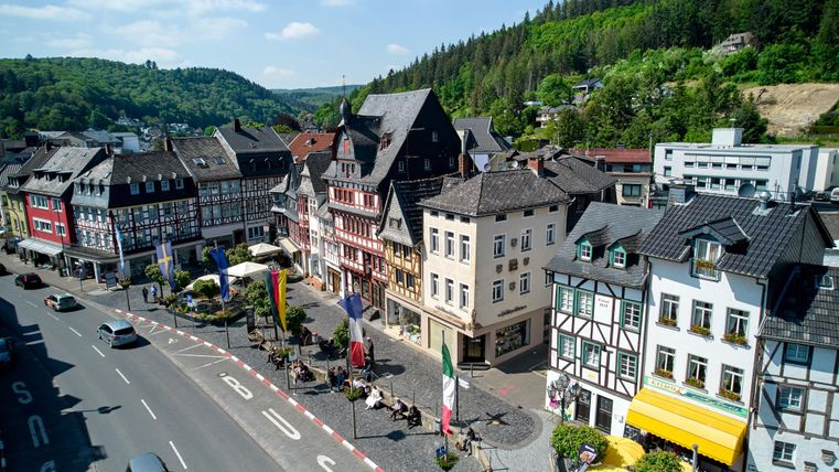 A picturesque town with traditional architecture and a green backdrop. The street is lively and lined with trees.