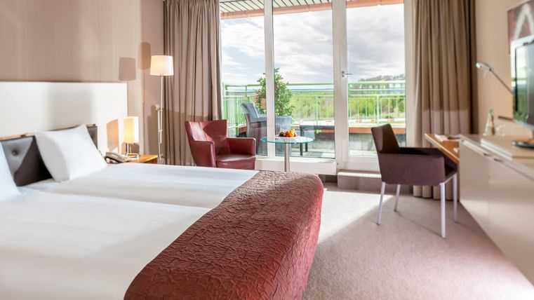 A modern hotel room with two beds and a view of a balcony. The room is bright and inviting, with stylish furniture and plenty of natural light.