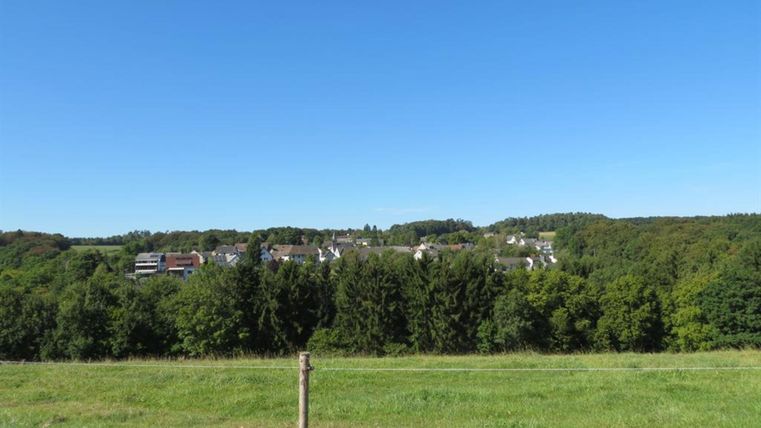 Eine malerische Landschaft mit einem klaren blauen Himmel. Im Hintergrund sind Häuser und viel grüne Vegetation zu sehen.