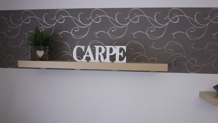 Een eenvoudige muur met een decoratieve witte tekst die "CARPE" zegt. Daaronder staat een plank met een kleine plant in een glas.