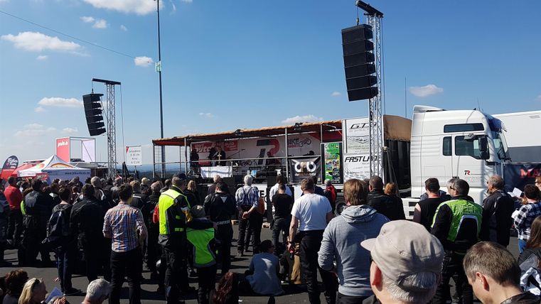 Eine große Menschenmenge versammelt sich vor einer Bühne mit Lautsprechern. Im Hintergrund sind ein Lkw und ein blauer Himmel mit einigen Wolken zu sehen.