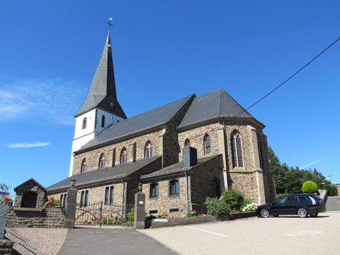 Eine schöne Kirche mit einem hohen Turm. Im Vordergrund sind ein Parkplatz und grünes Laub.