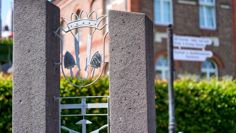 Een decoratieve metalen omheining met een wapen en plantmotieven. Op de achtergrond zijn ramen en wegwijzers zichtbaar.