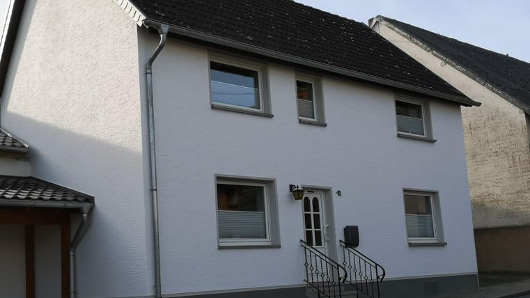 A modern, white residential building with a sloping roof. The building has several windows and an entrance door with stairs.