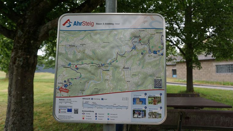 A map of the AhrSteig with marked hiking trails and points of interest. In the background, trees and a seating area can be seen.