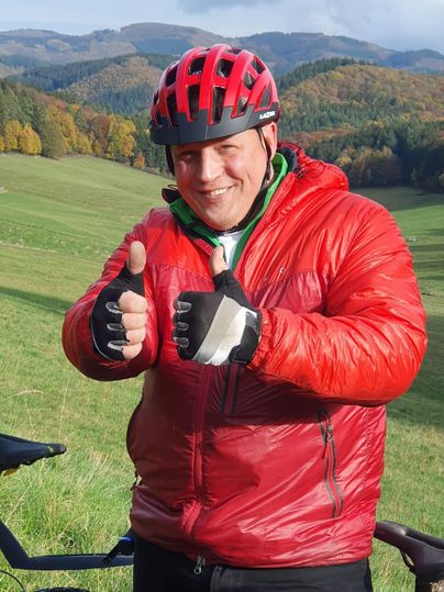 Ein Mann in einem roten Fahrradhelm und einer roten Jacke gibt zwei Daumen hoch. Im Hintergrund sind grüne Hügel und Bäume zu sehen.