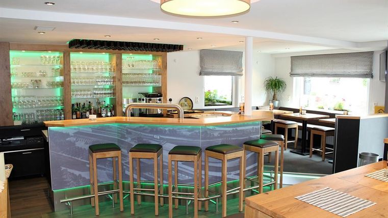 A modern bar with a wooden counter and green lighting. There are wooden chairs and inviting tables in the background.