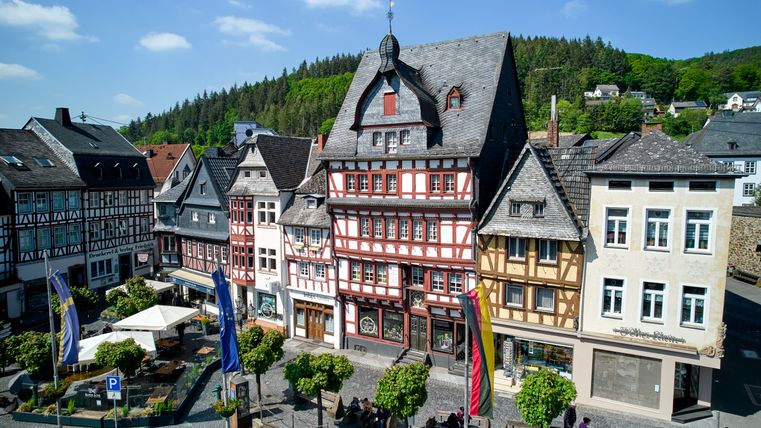 Historische Fachwerkhäuser am Marktplatz in Adenau, umgeben von Bäumen und Flaggen, bei sonnigem Wetter.