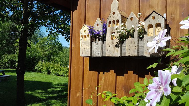 Eine dekorative Vogelhaus-Anordnung an einer Holzwand mit bunten Blumen. Im Hintergrund sieht man eine grüne Wiese und Bäume.