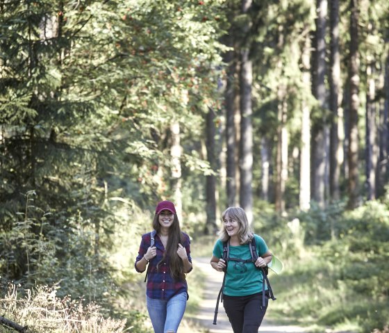 Zwei Frauen wandern fr&ouml;hlich auf einem Waldweg in der Hocheifel. Sie tragen Rucks&auml;cke und sind von hohen B&auml;umen umgeben., &copy; TI Hocheifel-N&uuml;rburgring,Jonathan_Andrews