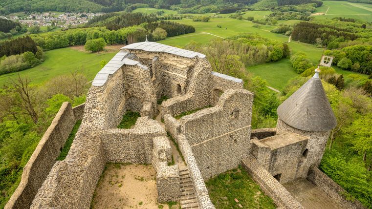 Een oude kasteelruïne met stenen en een ronde torenspits. Het omringende landschap is groen en heuvelachtig.