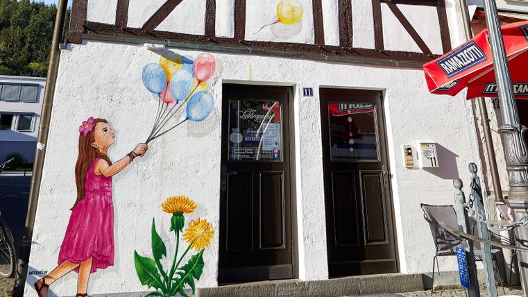 A wall art piece on a building showcases a small girl in a pink dress playing with colorful balloons. Next to the girl, yellow flowers are depicted.