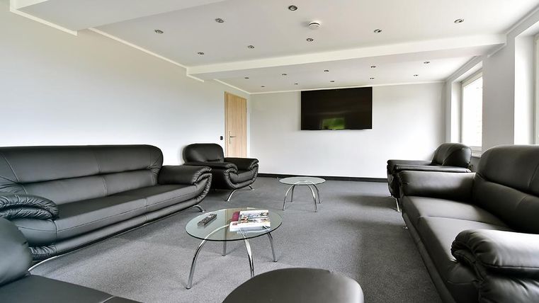 Ein modernes Wohnzimmer mit schwarzen Ledersofas und einem Glastisch. An der Wand hängt ein Fernseher, und große Fenster lassen viel Tageslicht herein.