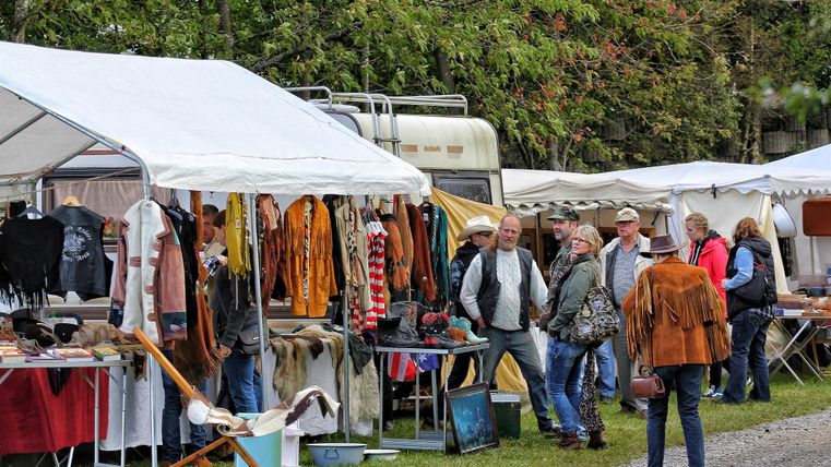 Ein bunter Markt mit mehreren Ständen, die Kleidung und Accessoires anbieten. Menschen schlendern zwischen den Ständen und betrachten die Ware.