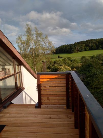 A wooden balcony with a beautiful view of green meadows and trees. The sky is cloudy, creating a peaceful atmosphere.