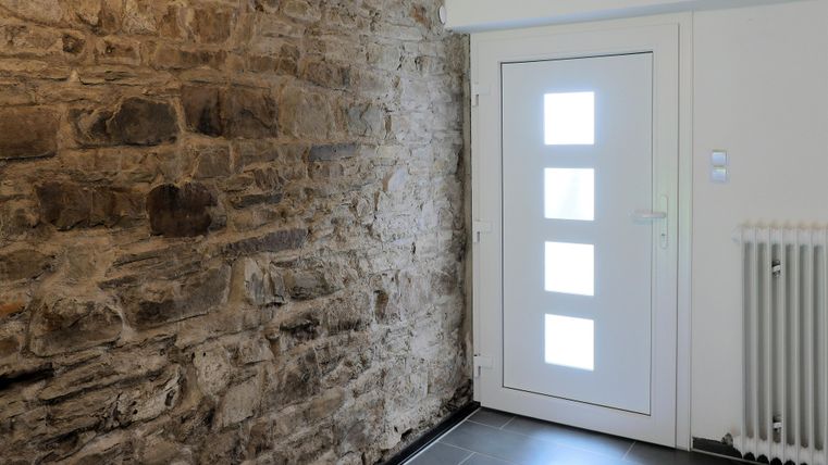 Ein moderner Eingangsbereich mit einer weißen Tür und einem Steiwand. Der Boden ist mit dunklen Fliesen ausgelegt.