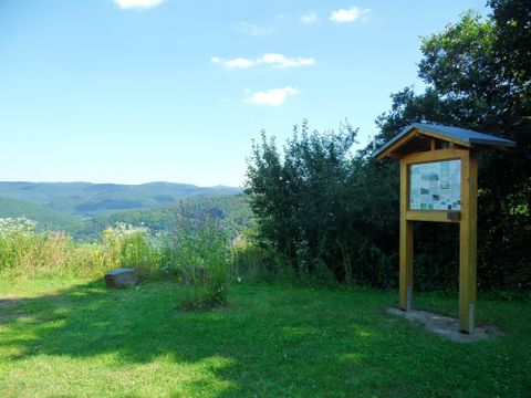 A beautiful view of green hills and a blue sky. In the foreground stands an information sign surrounded by grass and bushes.