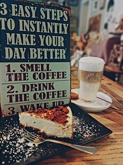 Un morceau de cheesecake sur une assiette à côté d'un verre de café. En arrière-plan, il y a un panneau avec les étapes pour améliorer la journée.