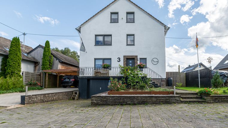 Ein großes, weißes Wohnhaus mit mehreren Fenstern und einem Balkon. Vor dem Haus gibt es einen gepflasterten Hof und einige grüne Pflanzen.