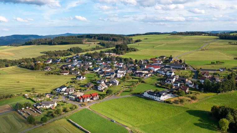 A picturesque landscape with a small village surrounded by green fields. Gentle hills and blue sky create a serene atmosphere.