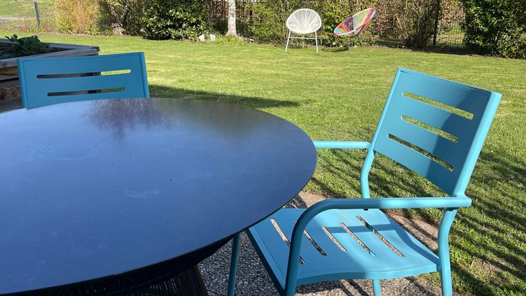Un beau jardin avec une table ronde et des chaises bleues. En arrière-plan, il y a des arbres et un coin salon qui a l'air confortable pour se détendre.