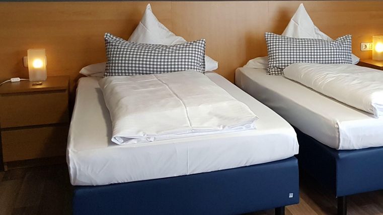 A modern bedroom with two single beds and cozy pillows. The walls are kept in a simple color tone, and there is a bedside lamp on each nightstand.