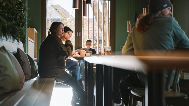 Eine gemütliche Café-Szene mit mehreren Gästen, die an Holztischen sitzen. Das Licht strömt durch große Fenster und schafft eine einladende Atmosphäre.