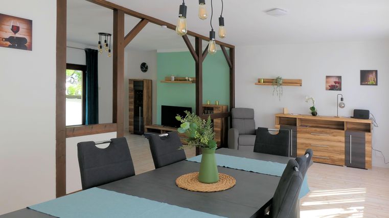 Ein modernes Wohnzimmer mit einem Esstisch, Stühlen und einer dekorativen Pflanze in der Mitte. Die Wände sind hell gestrichen und es gibt einige dekorative Elemente an den Wänden.