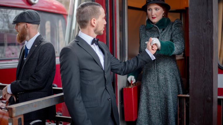 A man in a suit is shaking hands with a woman in a winter coat while they stand by a red train. Another person in a hat and suit is standing next to them.