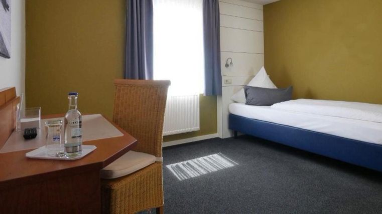 A simple hotel room with a single bed and a small table. It has bright walls and a large window that brings light into the room.