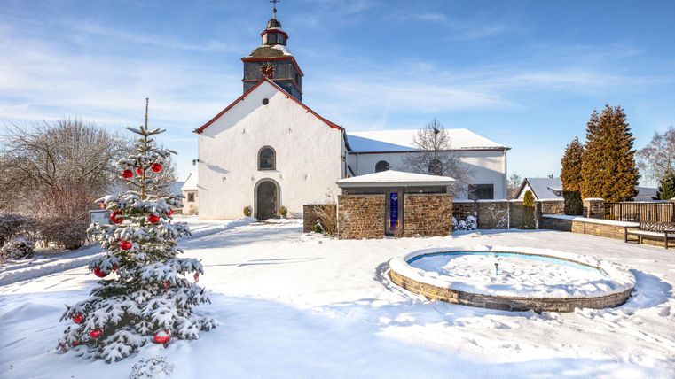 Eine verschneite Landschaft mit einer Kirche im Hintergrund. Im Vordergrund steht ein Weihnachtsbaum und ein Brunnen.