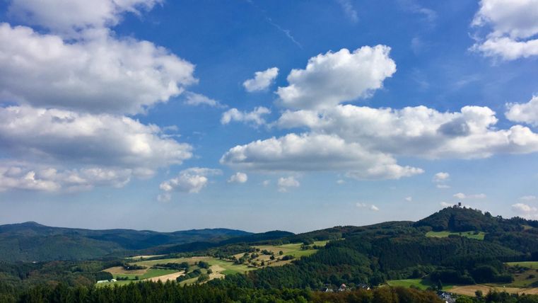 Landschaft mit Burgruine Nürburg auf einem Hügel, umgeben von Wäldern und Feldern unter blauem Himmel mit Wolken.