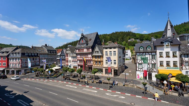 Eine malerische Stadtansicht mit historischen Fachwerkhäusern und einer grünen Hügelkulisse. Die Straße ist links sichtbar, und es gibt Schirme und Tische für Außenbewirtung.