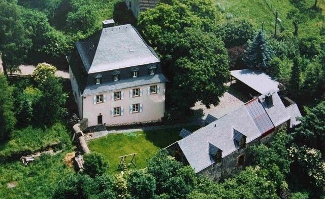 Ein schönes, zweigeschossiges Gebäude umgeben von Bäumen und einem grünen Garten. Im Hintergrund sind weitere kleine Gebäude und ein Spielplatz zu sehen.
