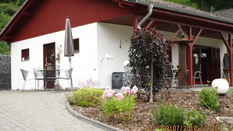 Une maison moderne avec un toit rouge et une terrasse. Les environs sont joliment aménagés avec des fleurs et des plantes.