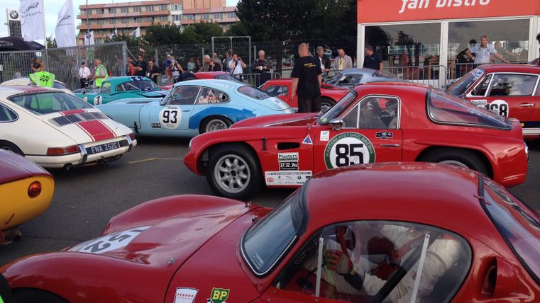 Eine Gruppe von klassischen Rennwagen steht auf einem Parkplatz. Im Hintergrund sind Zuschauer und ein Fan-Bistro zu sehen.