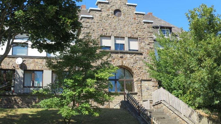 Ein historisches Gebäude aus Stein mit großen Fenstern. Um das Gebäude herum wachsen Bäume und es gibt eine Treppe, die zu einem flachen Gelände führt.