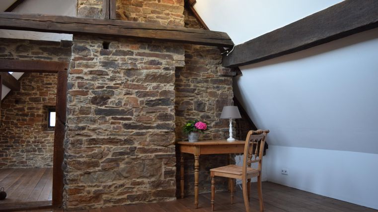 A cozy attic room with a stone wall and wooden beams. There is a lamp and a vase with flowers on a table.