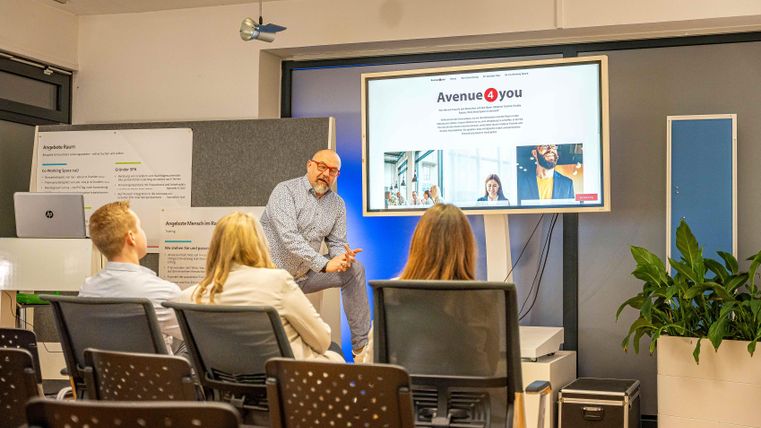 Ein Mann präsentiert vor einer Gruppe von Zuhörern in einem modernen Besprechungsraum. Auf dem Bildschirm wird die Website "Avenue 4 you" angezeigt.
