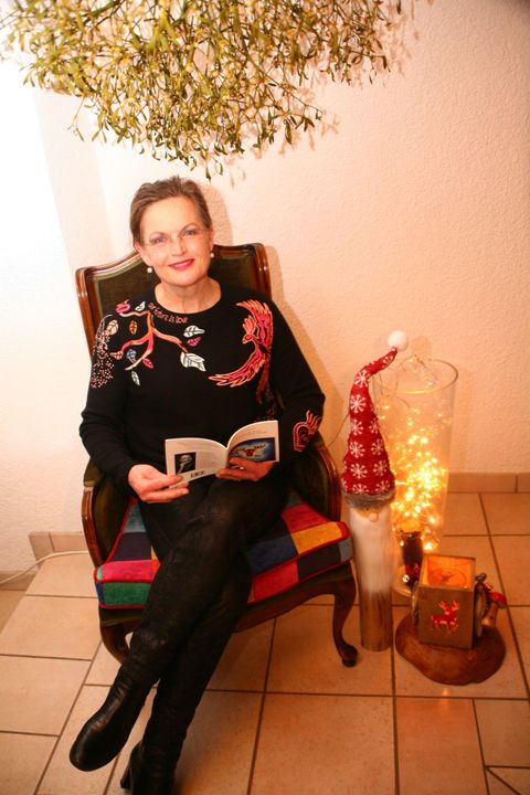 A woman is sitting comfortably in a chair, reading a book. Next to her is a decorative Christmas lantern and a small Christmas tree.