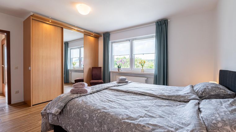 Een gezellige slaapkamer met een groot bed en een houten vloer. De ramen laten veel daglicht binnen en de ruimte is eenvoudig gedecoreerd.