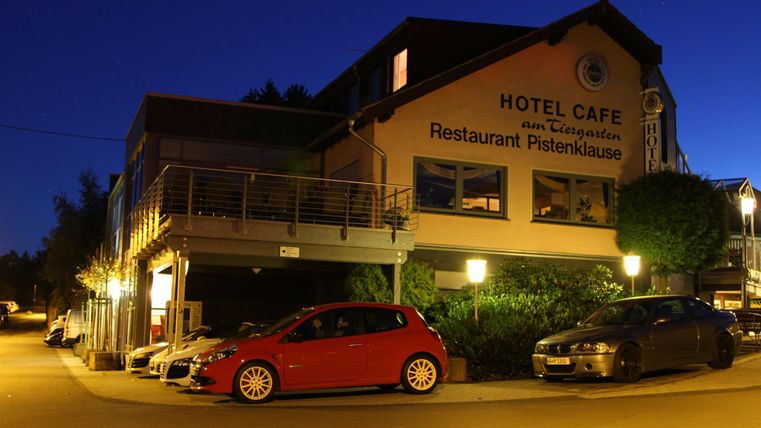 A building with the signage "Hotel Café" and "Restaurant Pistenklause" is illuminated at night. Cars are visible on the road in front of the hotel.