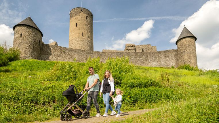 Een gezin met een kinderwagen staat op een pad voor een kasteel met torens. De omgeving is groen en zonnig.