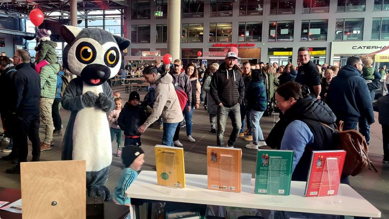 Ein überdimensionales Tierkostüm steht an einem Tisch mit mehreren bunten Büchern. Im Hintergrund ist eine Menschenmenge in einer belebten Umgebung zu sehen.