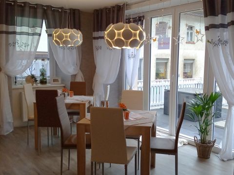 A modern dining area with a wooden table and chairs. Large windows provide plenty of daylight, while decorative lamps illuminate the space.